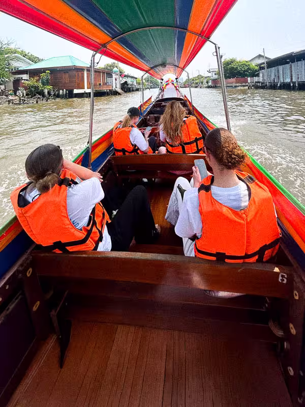 Four people wearing orange life jackets seated in a long-tail boat with a colorful canopy, traveling on a river with houses on the banks.