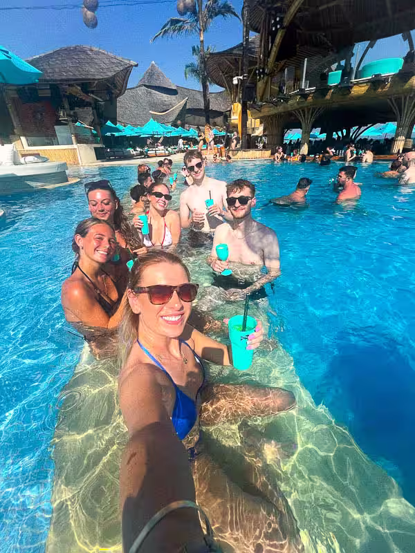 Group of smiling young adults in swimwear sitting in a pool at a tropical resort holding blue drinks.