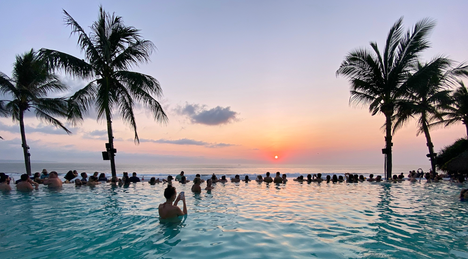 A group of people wallow at the edge of an infinity pool, surrounded by palm trees, taking pictures of the sunset.
