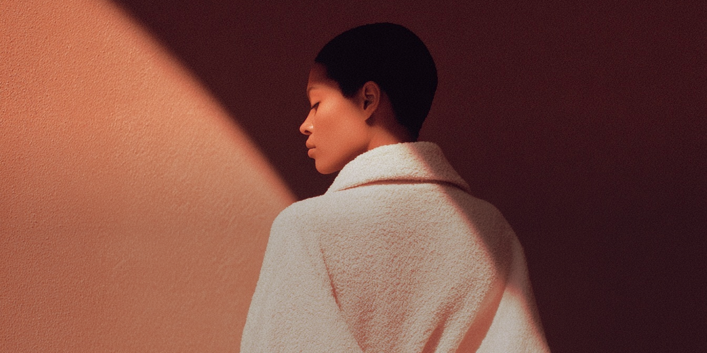 Profile of a person with short hair wearing a textured white coat, standing against a dark reddish-brown wall with a diagonal light beam.