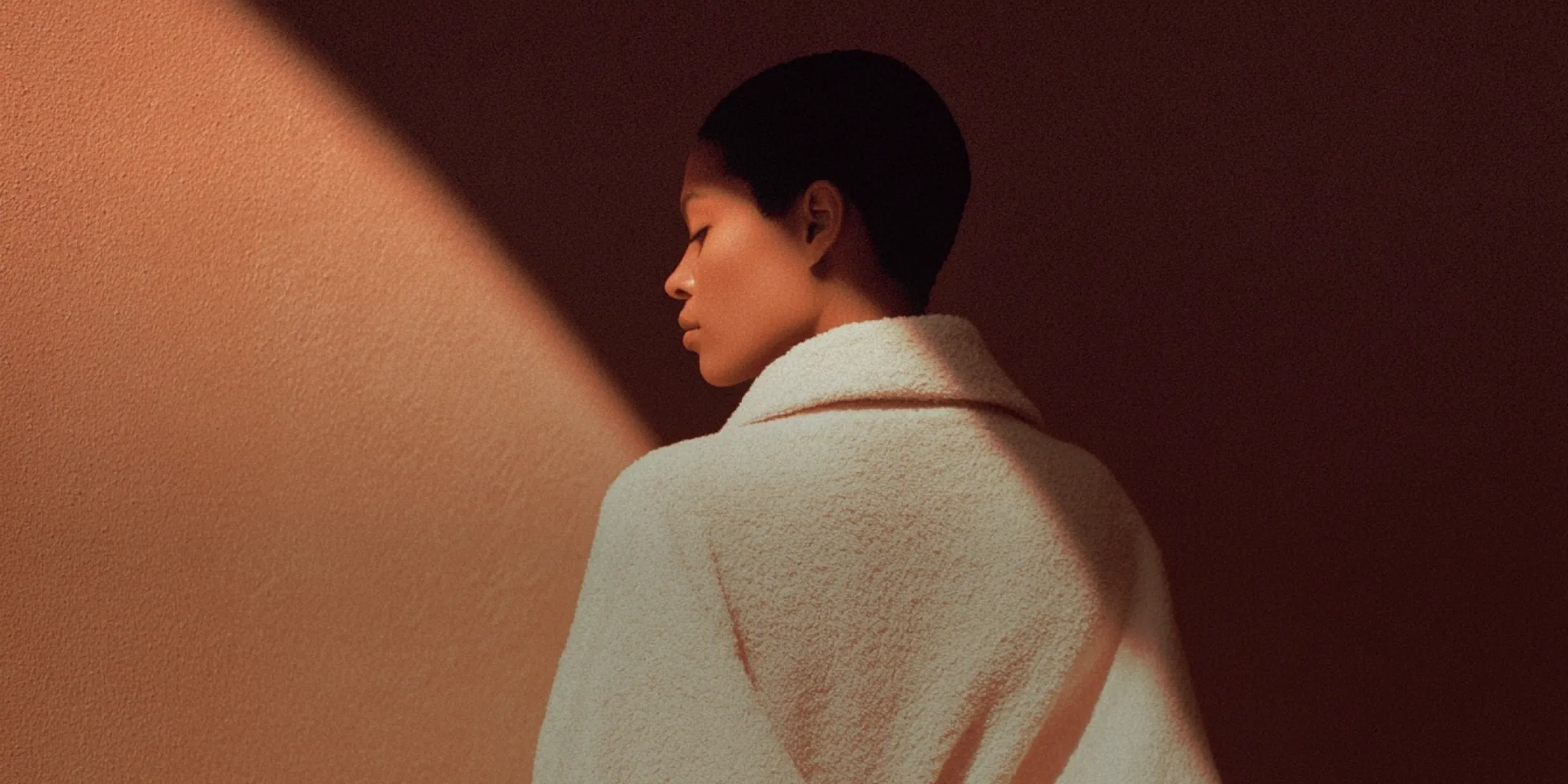 Profile of a person with short hair wearing a textured white coat, standing against a dark reddish-brown wall with a diagonal light beam.
