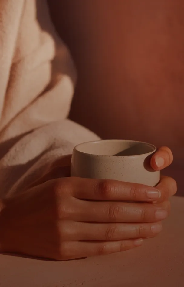 Woman in beige uniform preparing a stack of white towels on a massage table in a room with warm, angled sunlight and textured orange walls.