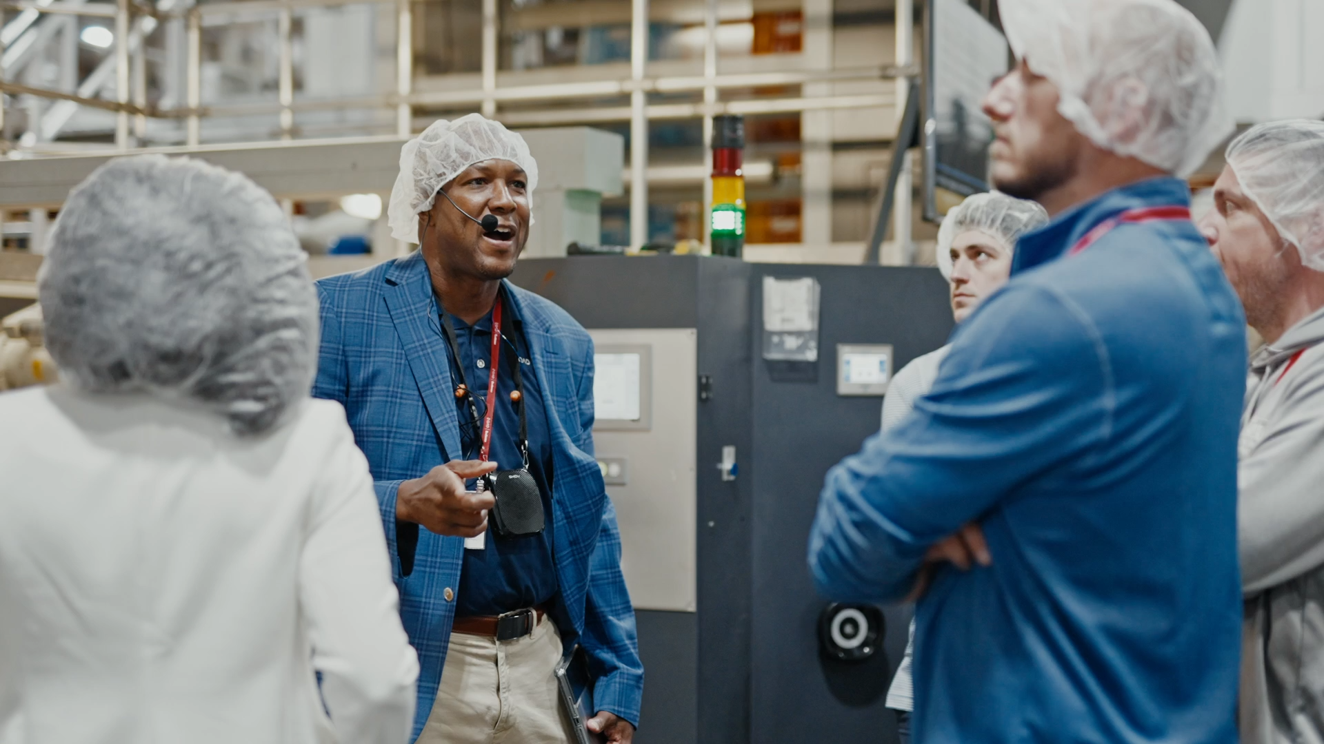 Factory manager in a blue blazer using a headset to lead a safety meeting with workers wearing hairnets in an industrial setting.
