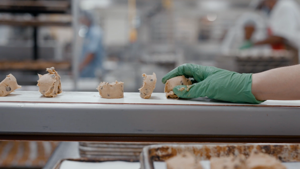 Gloved hand placing portions of cookie dough on a tray in a commercial bakery setting.