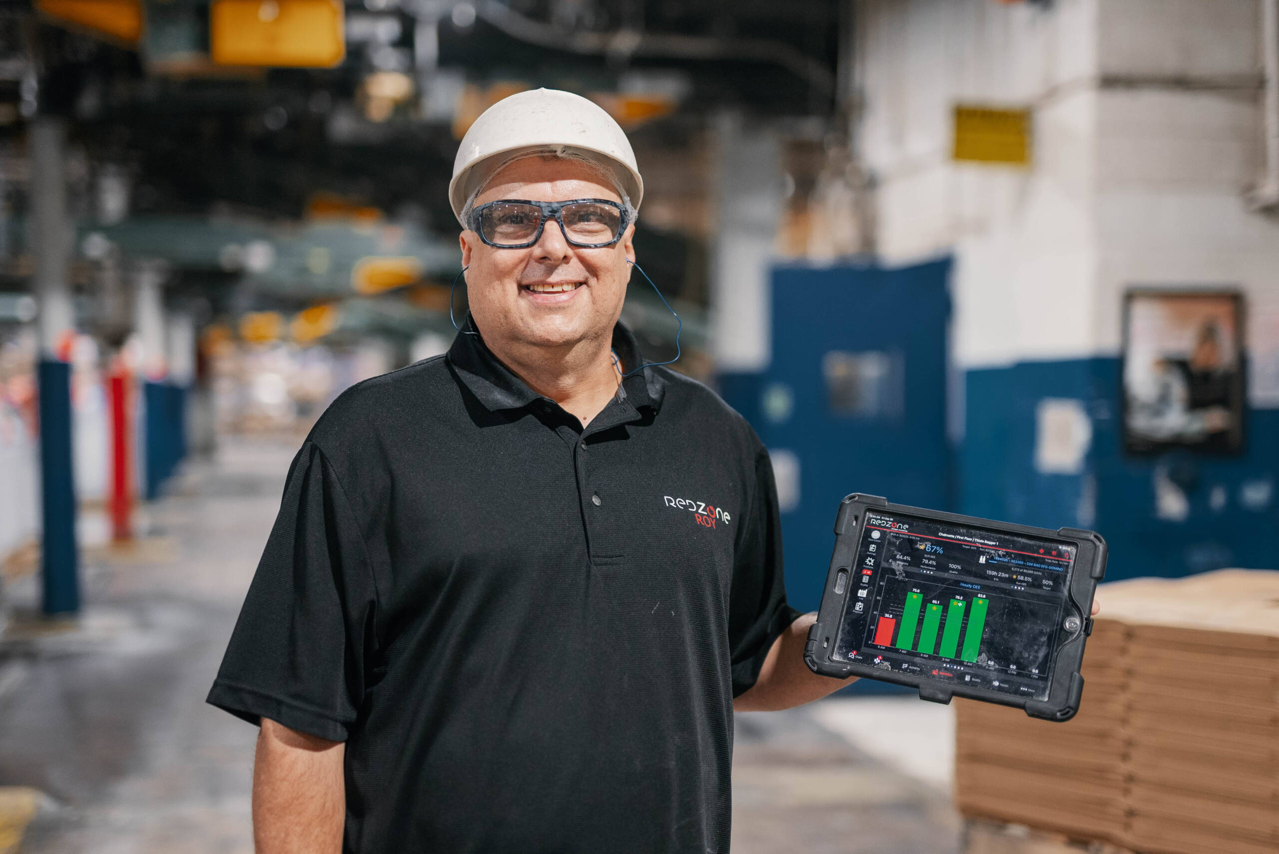 Man wearing a white hard hat and safety glasses holding a tablet displaying performance charts in an industrial warehouse.