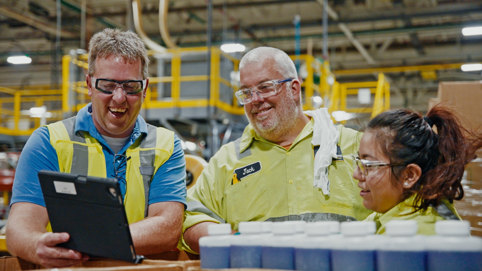 Three factory workers wearing safety glasses and high-visibility vests looking at a tablet and smiling.