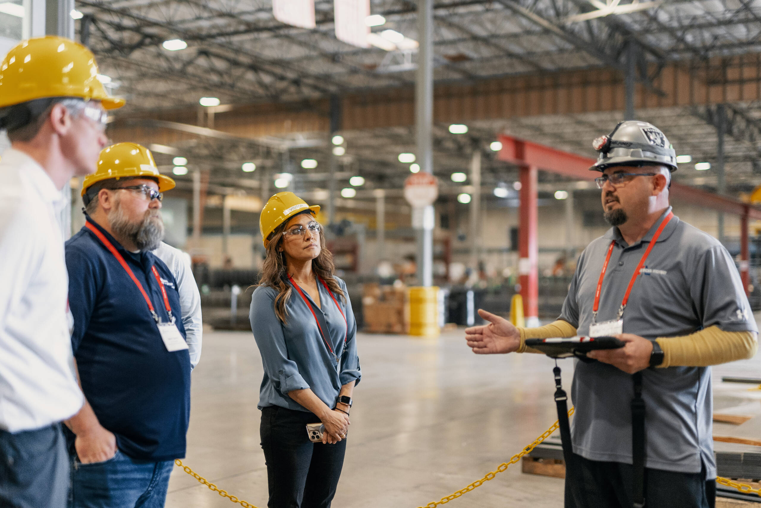 Group of construction workers wearing hard hats and safety glasses engaged in discussion inside a large industrial warehouse.