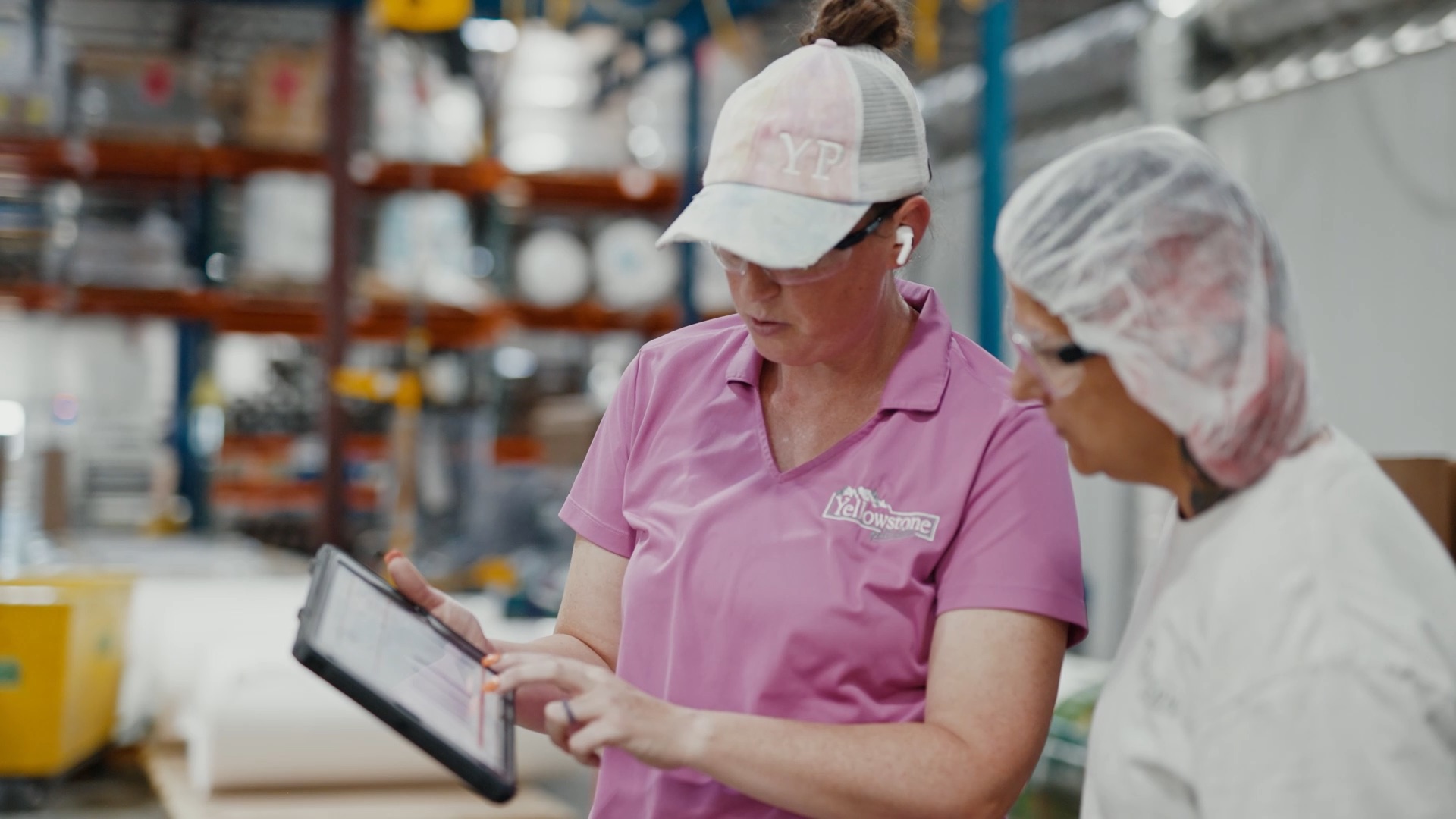 Two women in a warehouse reviewing information on a tablet, one wearing a pink Yellowstone shirt and cap.