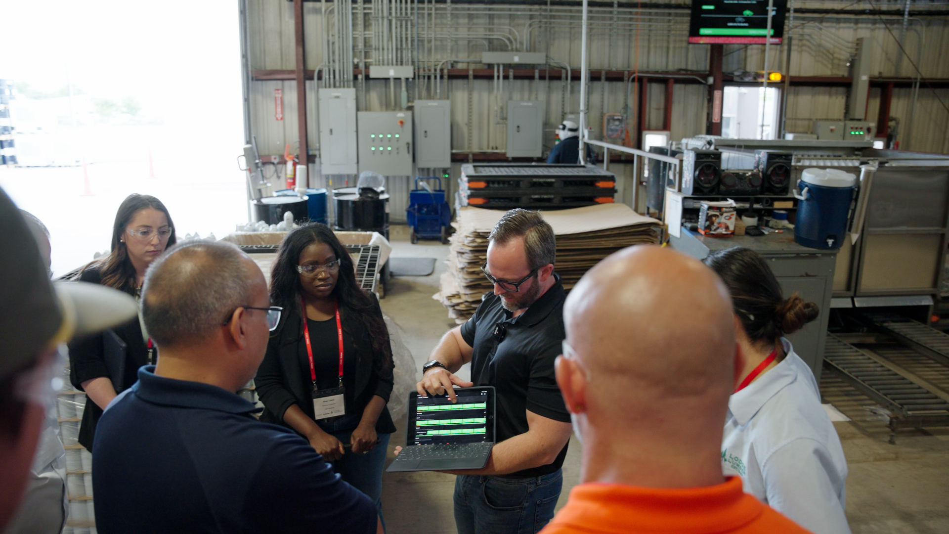 Man demonstrates data on a tablet to a group of workers wearing safety glasses in a warehouse.
