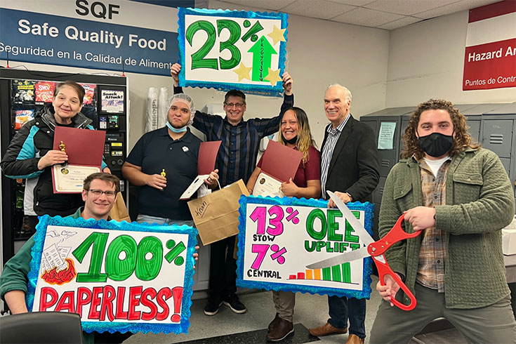 Six people in a workplace holding colorful signs showing achievements: one sign says 23% Productivity Up, another shows 100% Paperless, and the third displays 13% OEE Uplift and 7% Stov Central, with one person holding a large red scissors.
