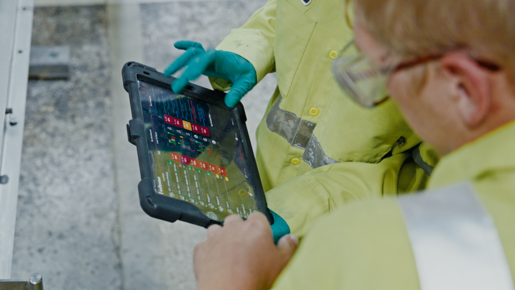 Two workers in yellow protective clothing and gloves looking at a rugged tablet displaying colorful data and graphs in an industrial setting.