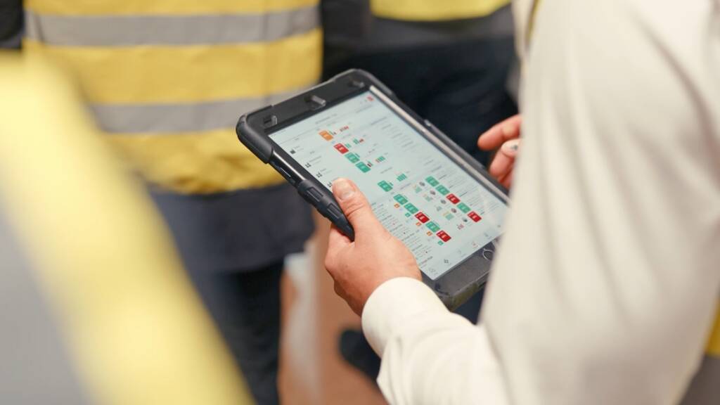 Person in a white shirt holding a rugged tablet displaying a digital dashboard with colored charts, with people in yellow safety vests in the background.