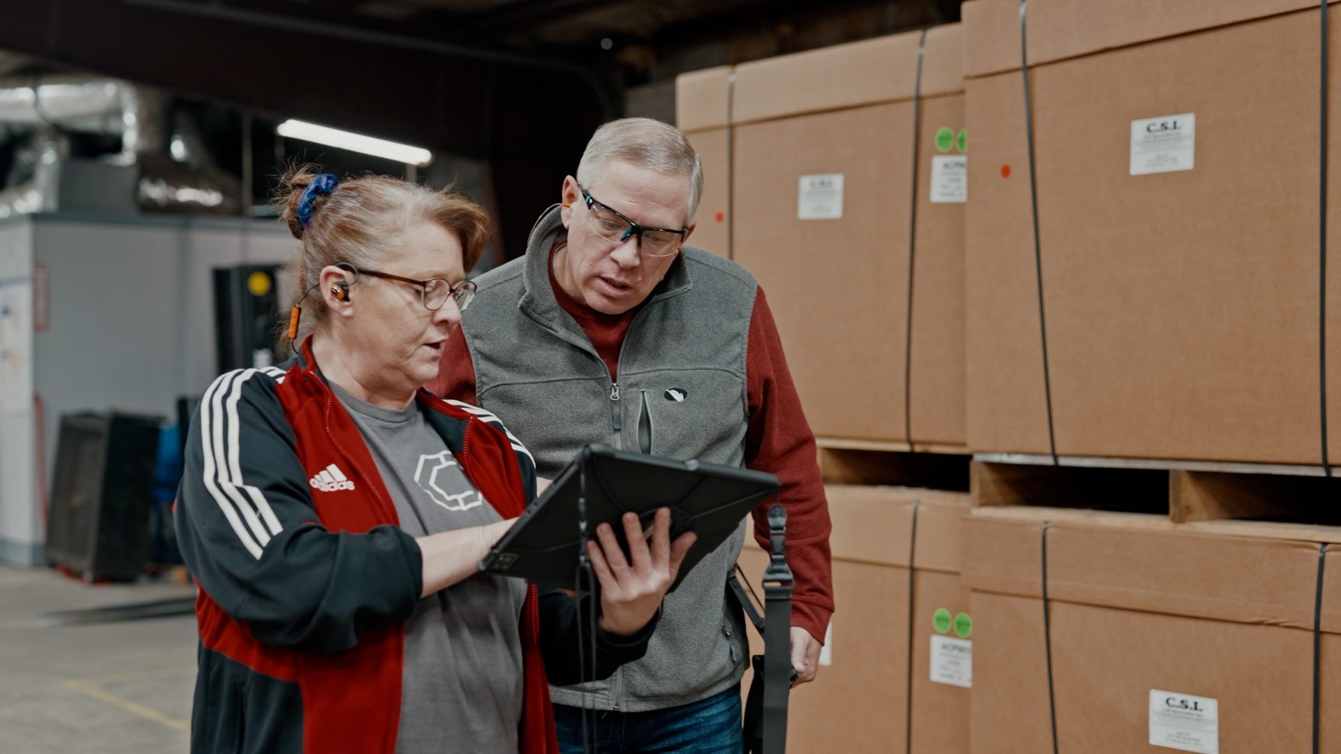 Two warehouse workers, a woman and a man, examining a tablet in front of stacked cardboard boxes on pallets.