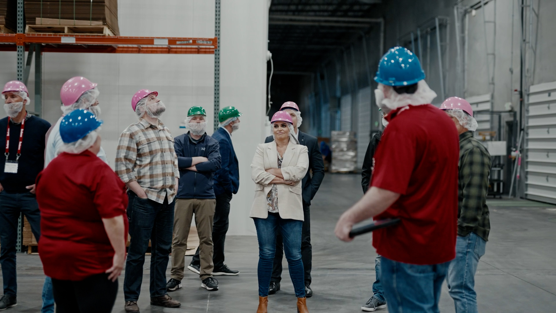 Group of workers wearing colored hard hats and hair nets in an industrial warehouse.