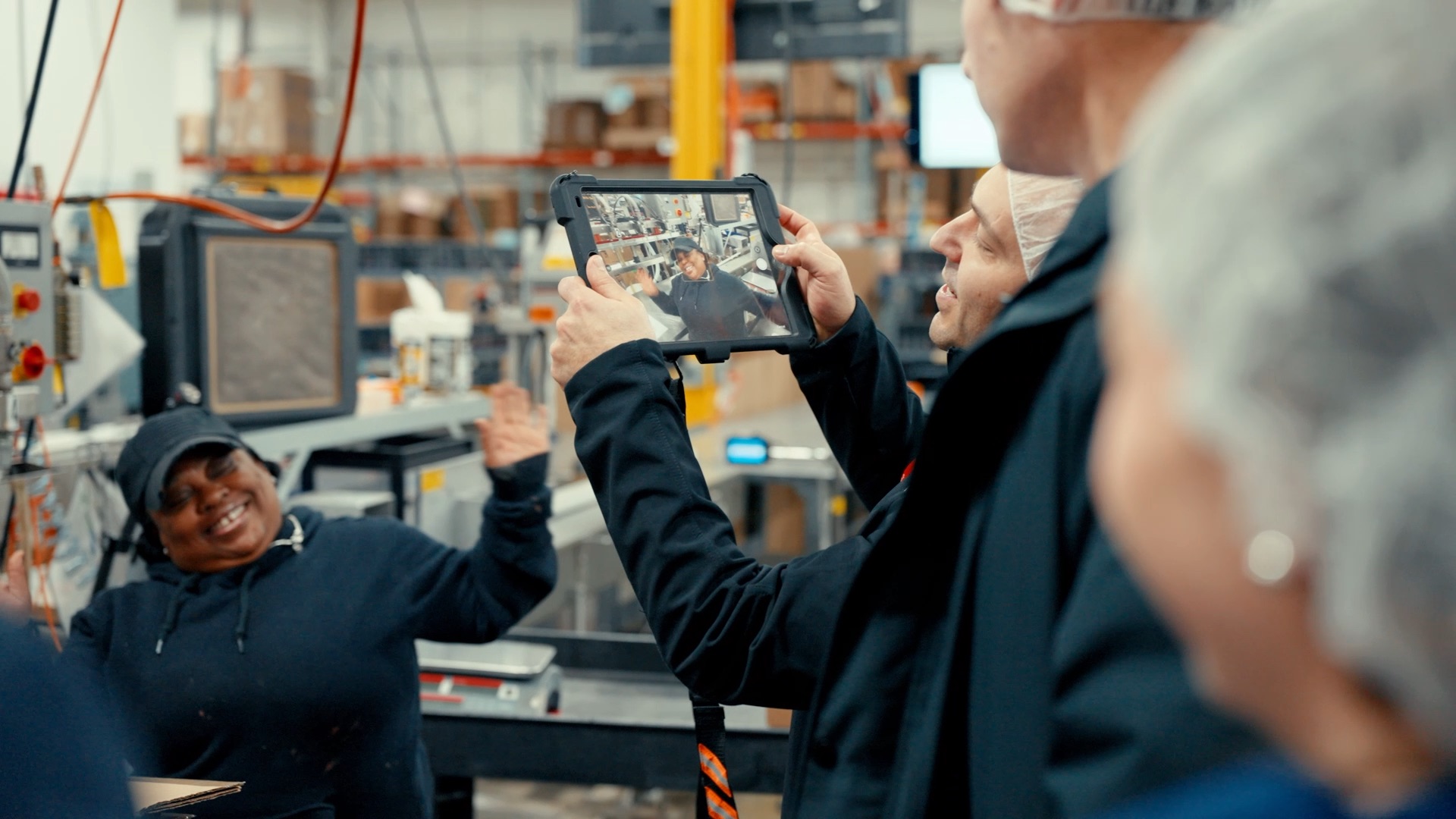 Factory worker smiling and waving during a video call displayed on a tablet held by a man in an industrial setting.