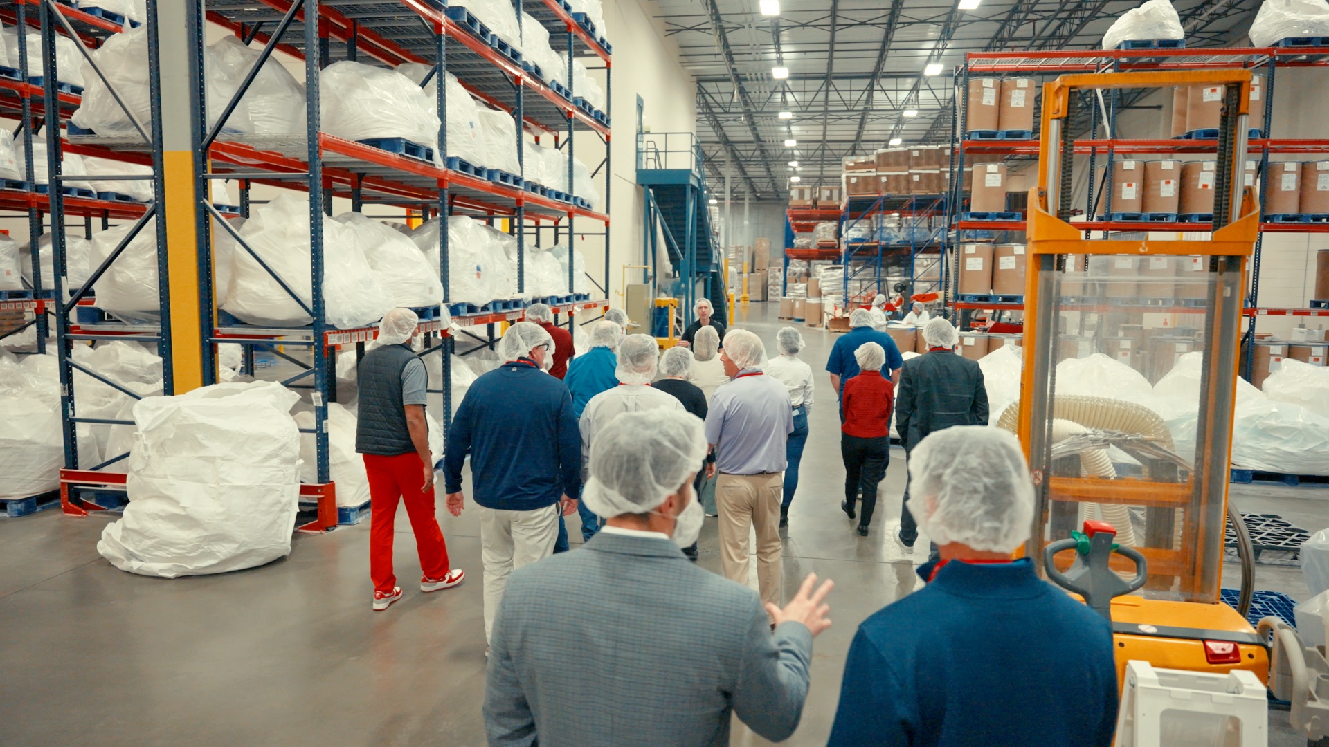 Group of people wearing hairnets touring a large warehouse filled with stacked pallets and packaging materials.