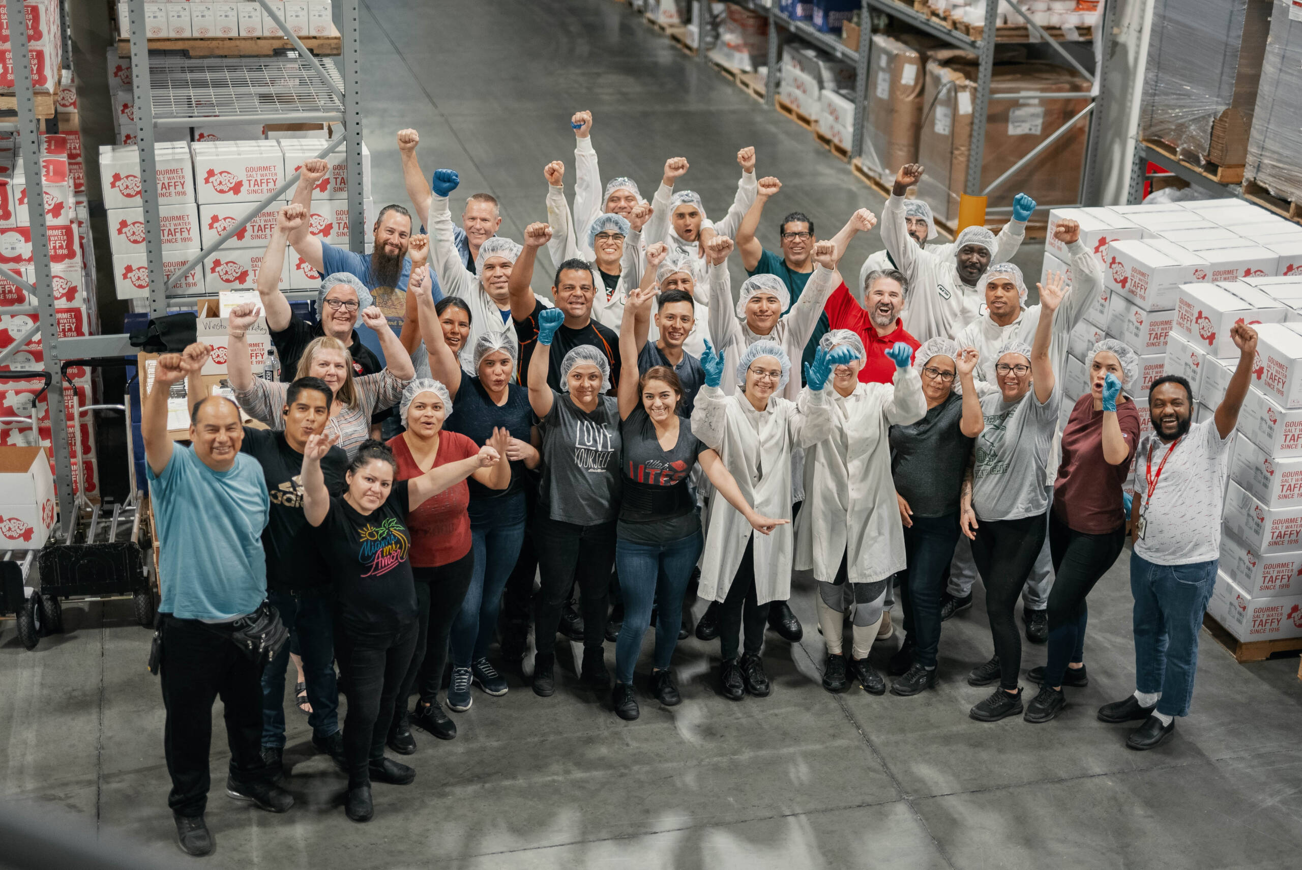 Group of diverse factory workers in a warehouse raising their fists and smiling surrounded by stacked boxes labeled Gourmet Salt Water Taffy.