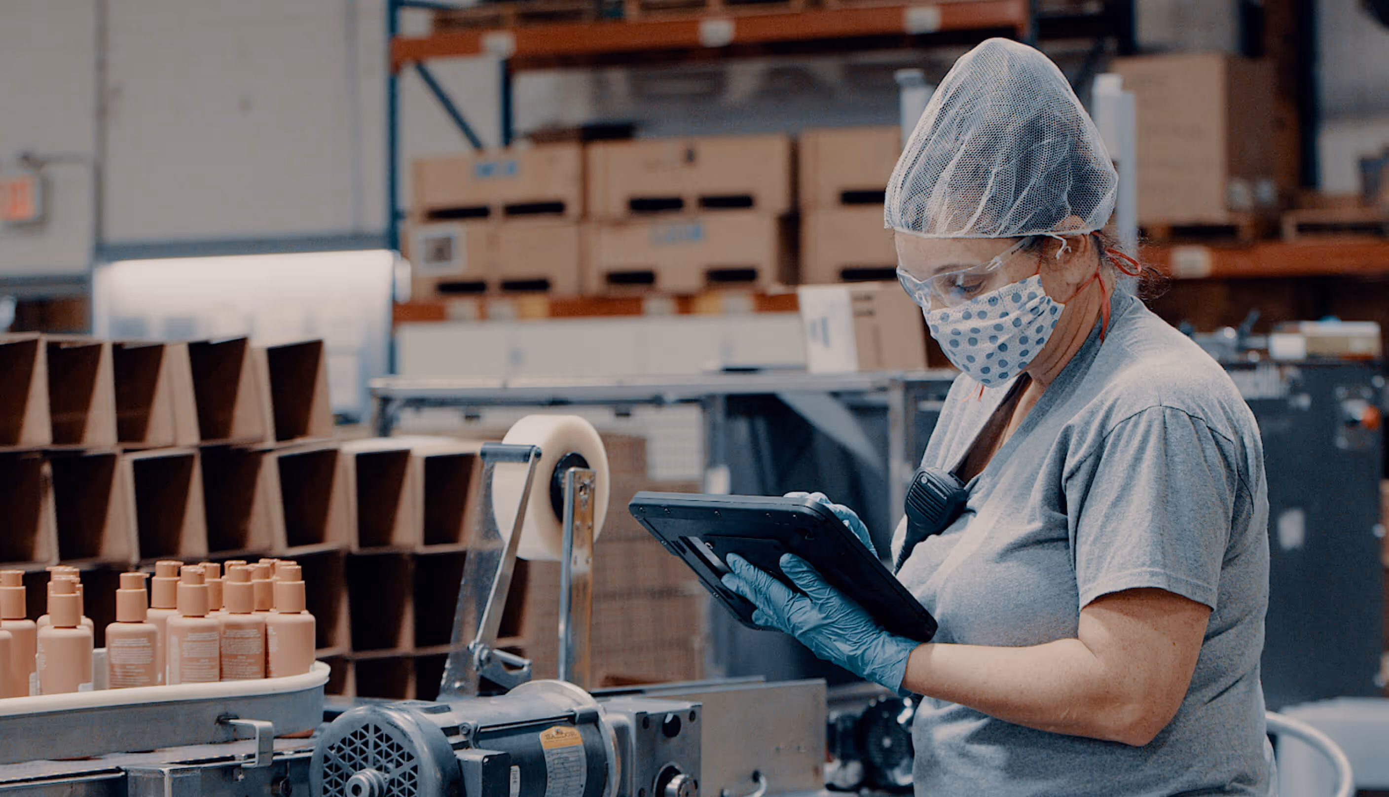 Factory worker wearing a mask, gloves, and hairnet uses a tablet while standing beside a conveyor belt with plastic bottles in a packaging facility.