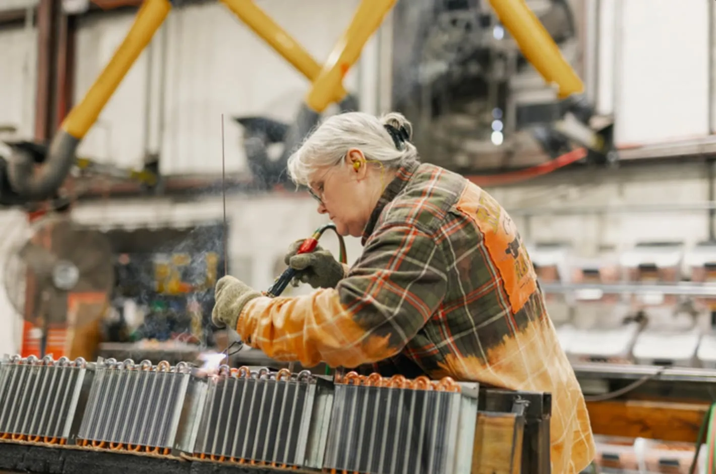Woman wearing safety glasses and earplugs using a welding torch on metal coils in an industrial workshop.