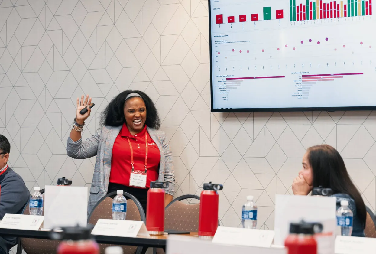 Woman in red shirt and gray blazer smiling and raising her hand while presenting data charts on a screen in a meeting room.