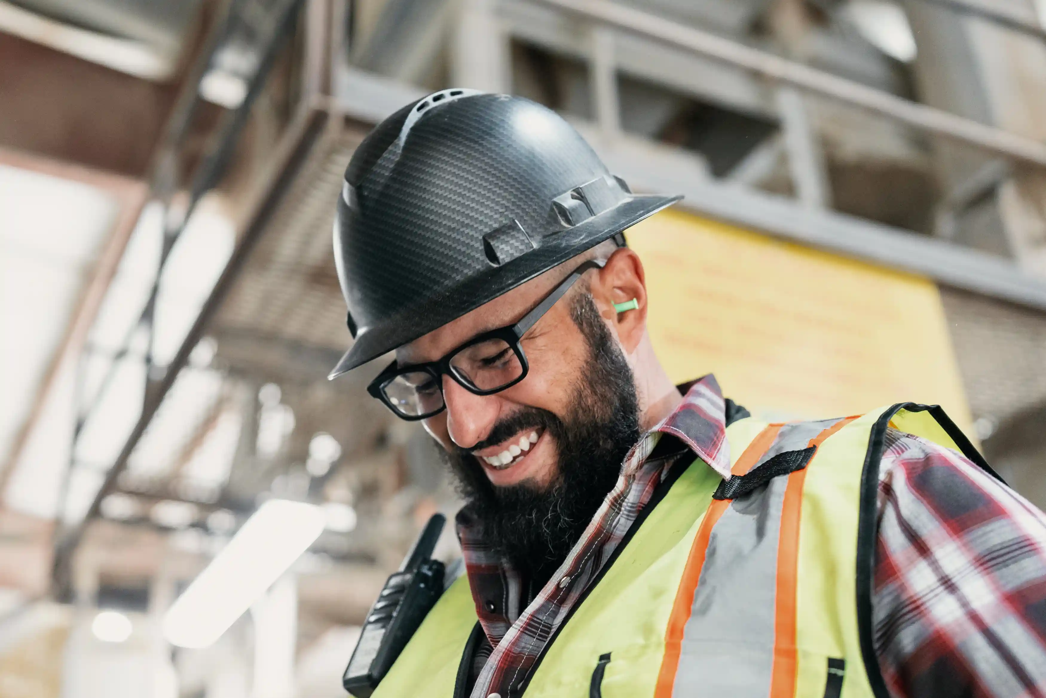 Smiling construction worker wearing a black hard hat, glasses, a high-visibility vest, and earplugs, indoors at a work site.
