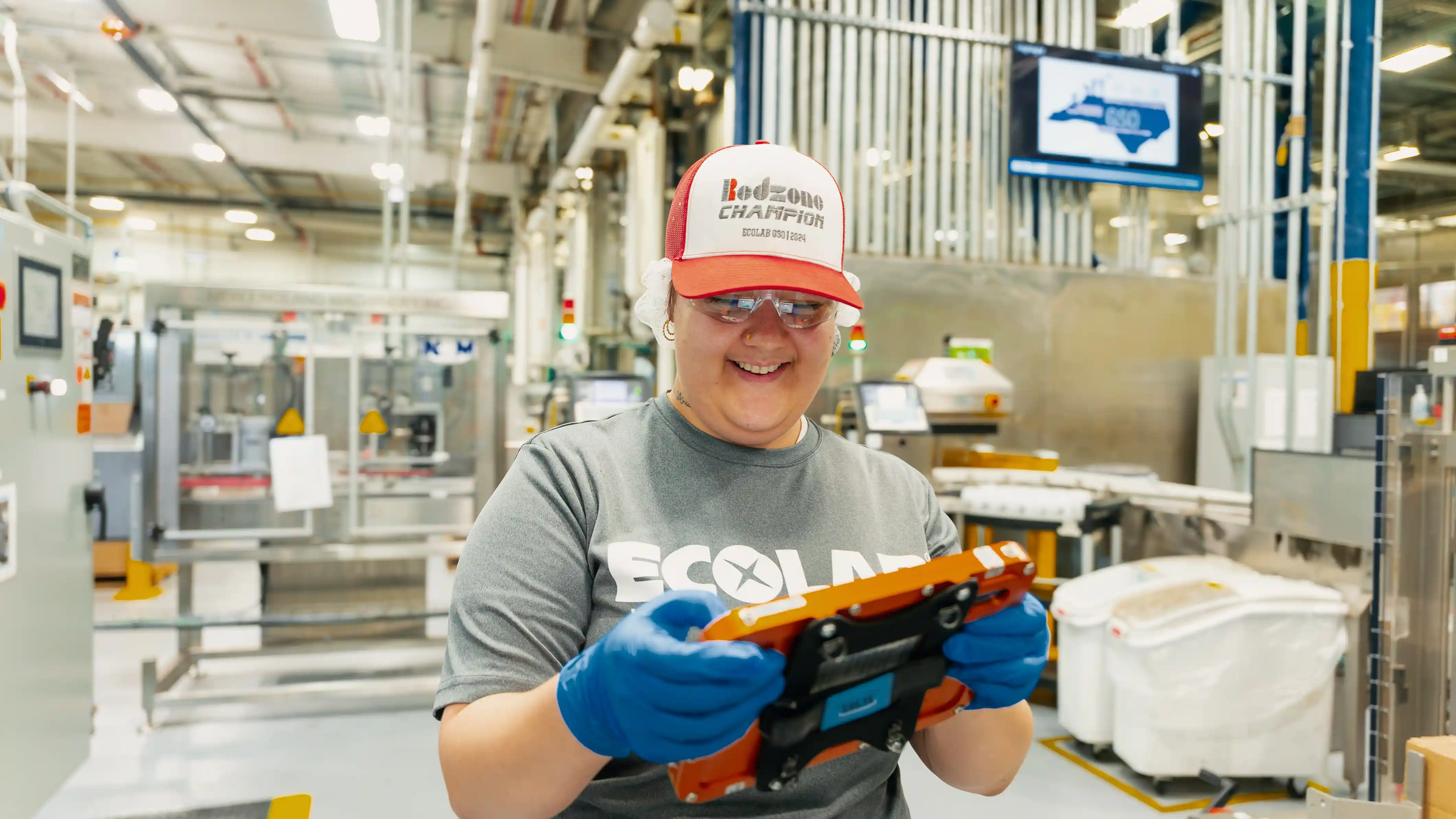 Smiling factory worker wearing a Redzone Champion cap, blue gloves, and safety glasses, holding an orange device in an industrial setting.