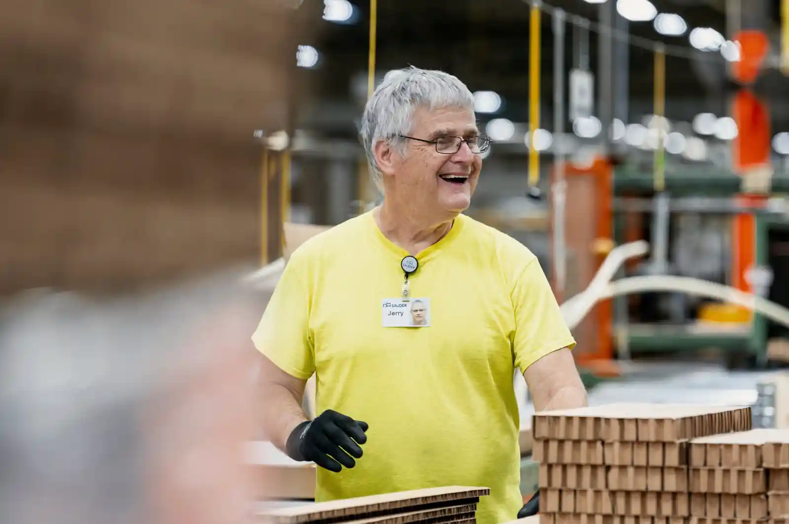 Smiling older man wearing a yellow shirt and black gloves working in a factory with stacks of cardboard.
