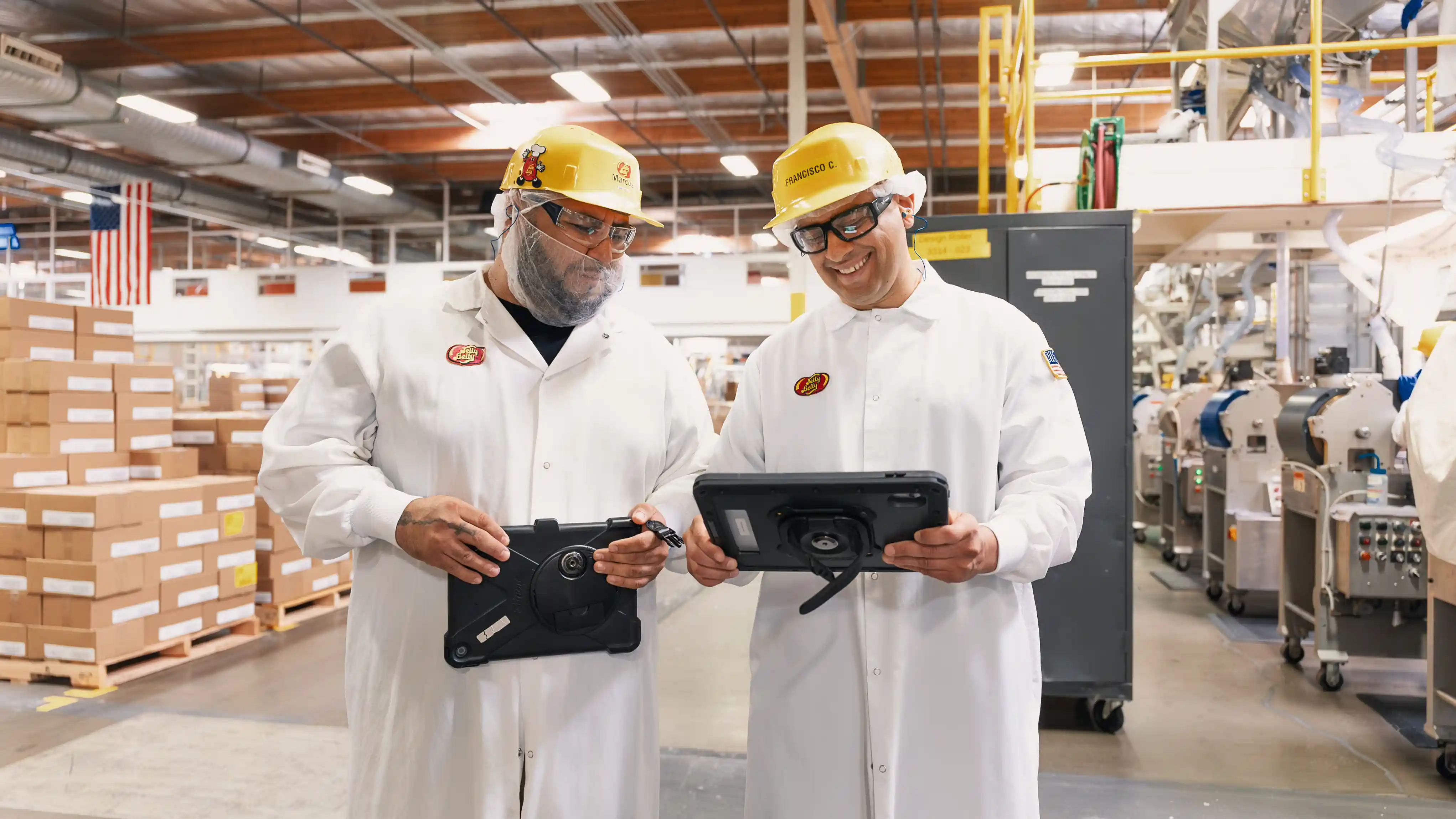Two factory workers in white coats and yellow hard hats holding tablets and smiling inside a manufacturing facility.