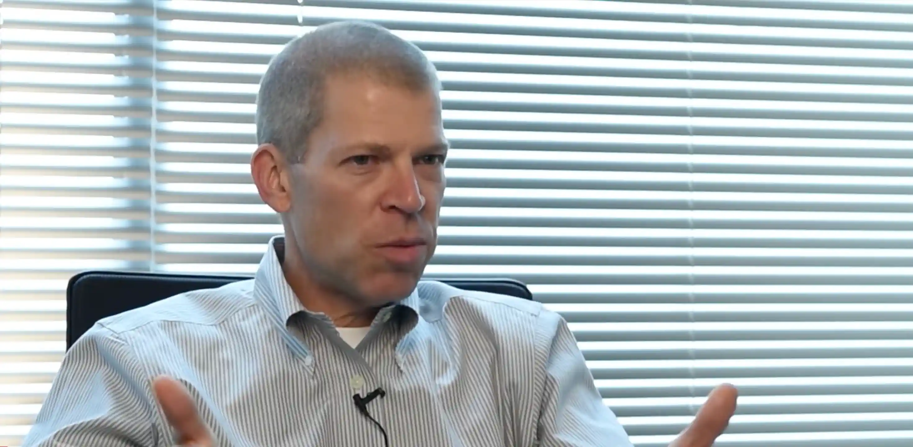 Man with short hair in a striped shirt speaking with hand gestures in front of horizontal window blinds.