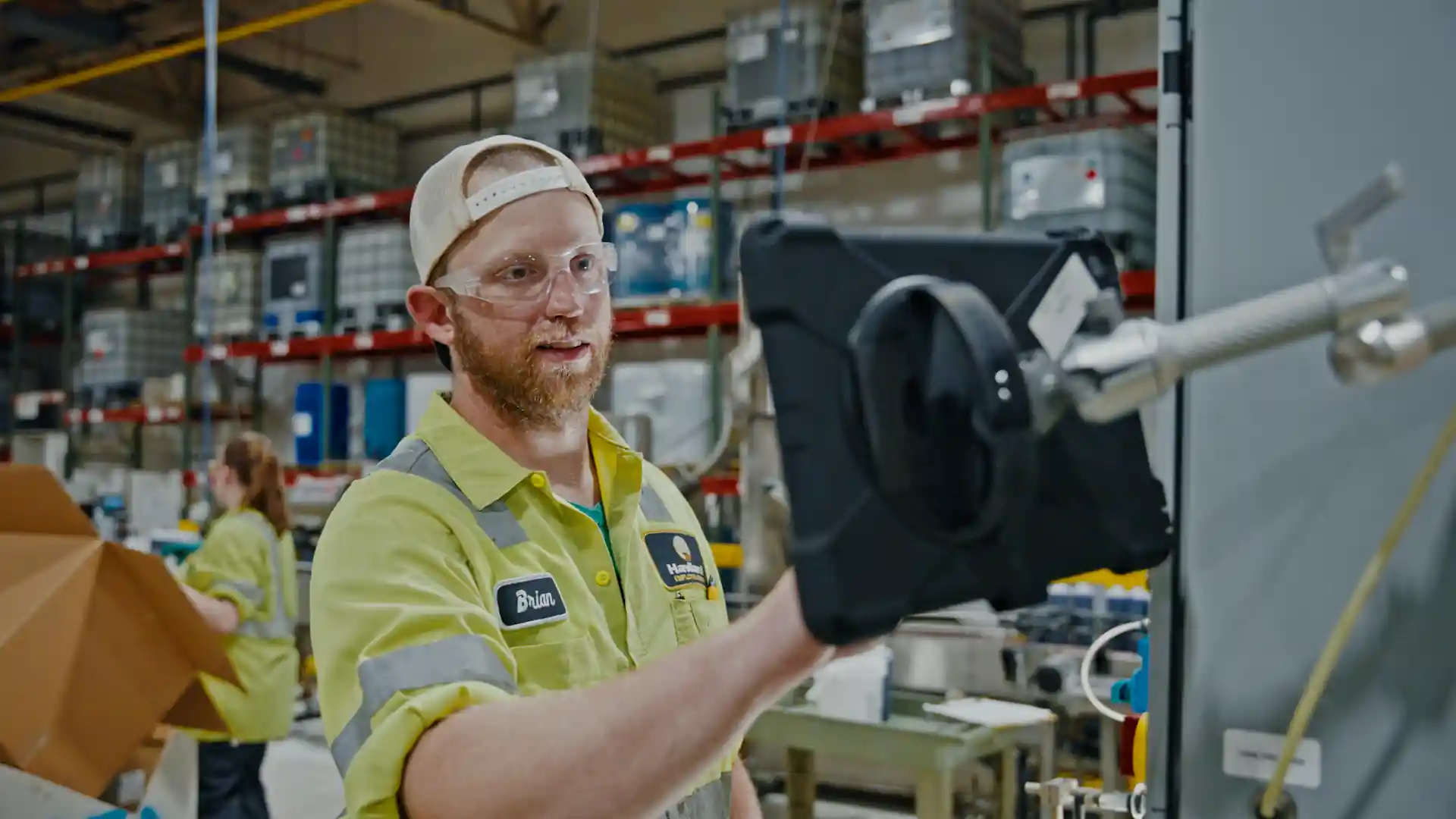A factory worker named Brian wearing safety glasses and a yellow uniform operates a machine using a touchscreen device in an industrial setting.