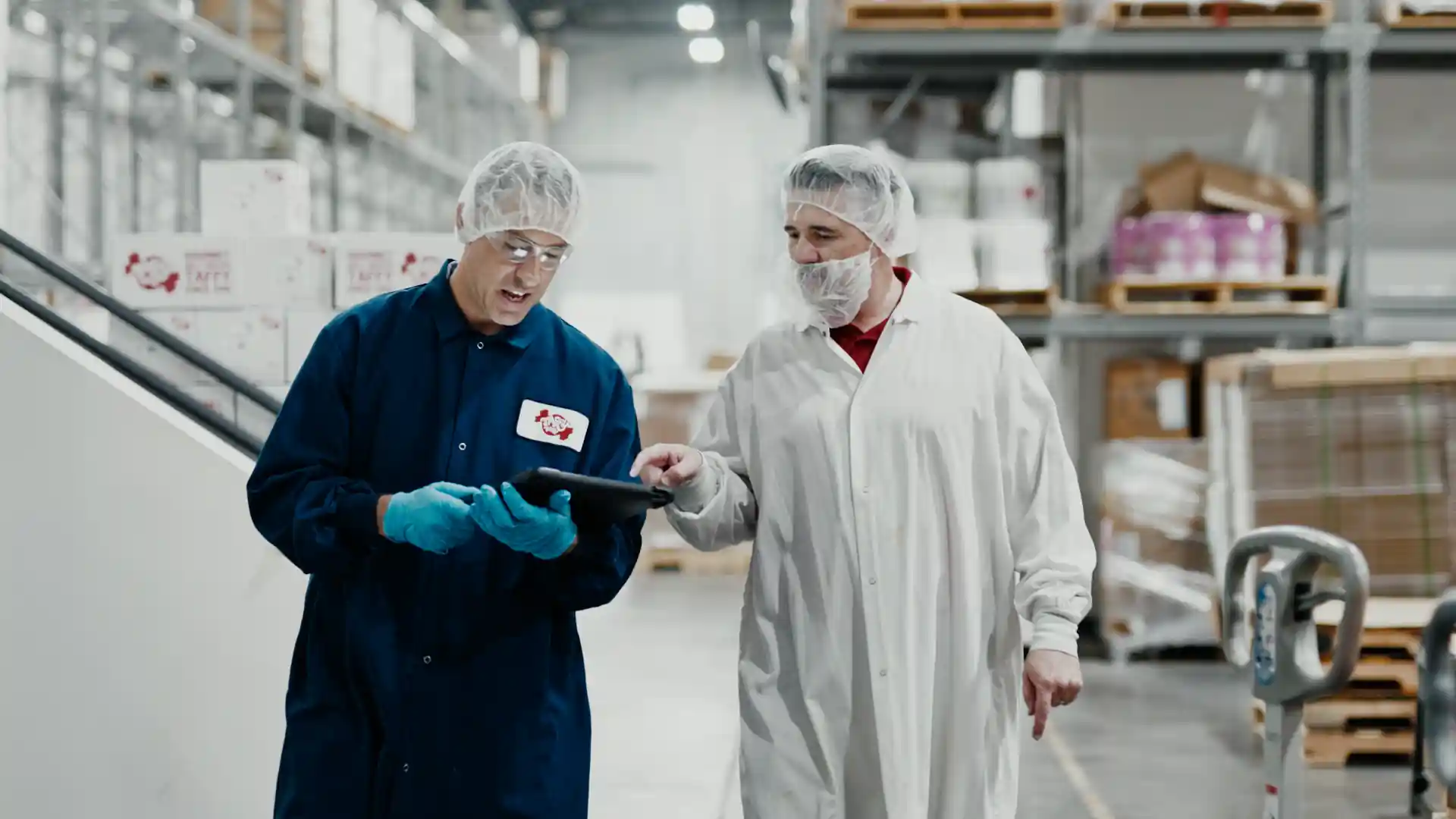 Two warehouse workers in protective clothing and hairnets discussing something on a tablet inside a storage facility.