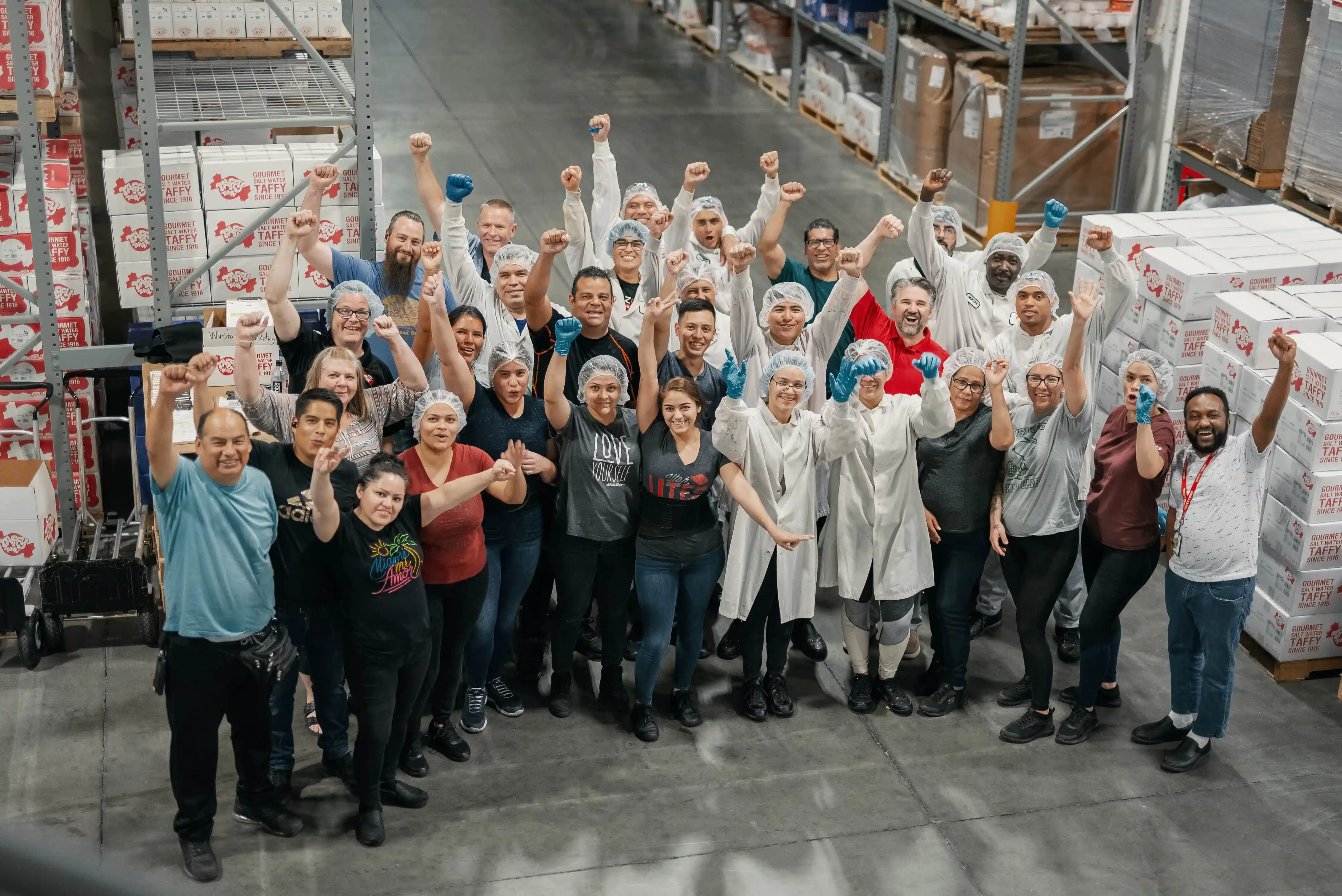 Group of factory workers in protective hairnets and gloves raising fists in celebration inside a warehouse.