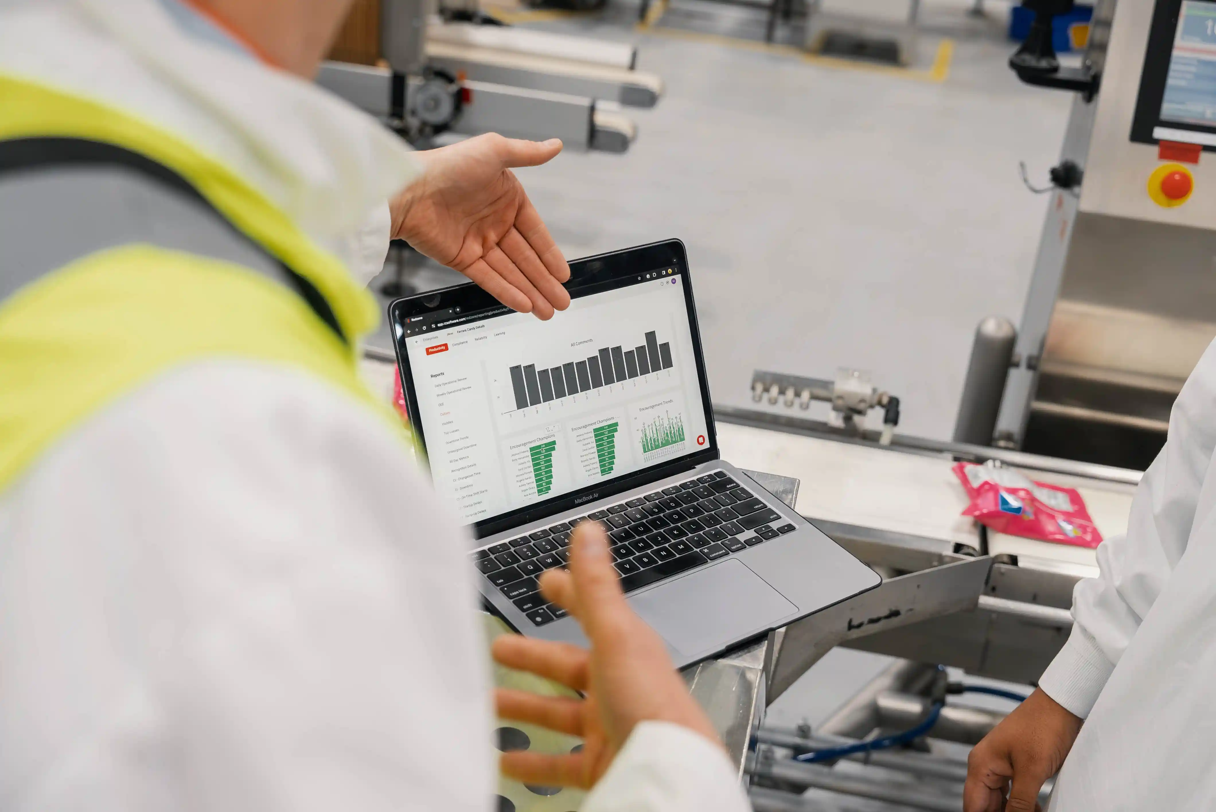 Two people in a factory discussing data charts on a laptop screen showing bar and engagement graphs.