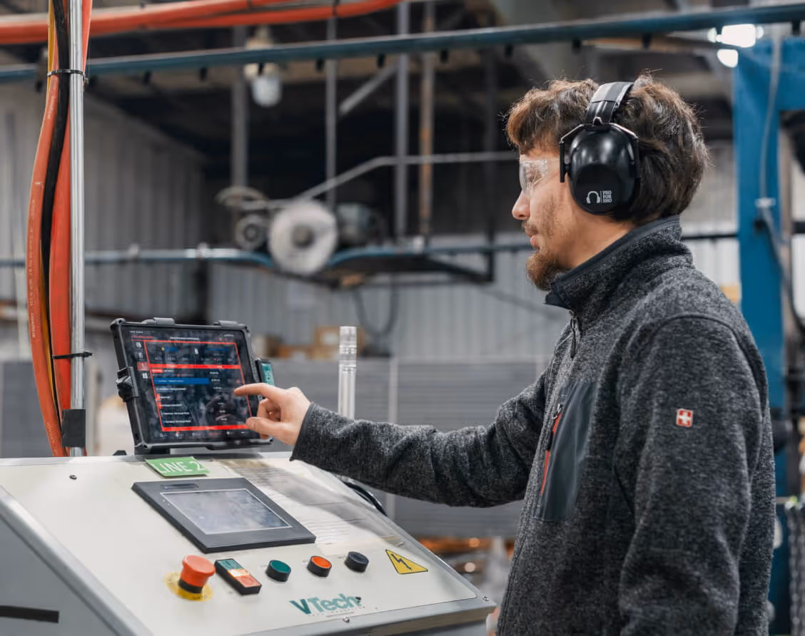 Man in safety glasses and earmuffs operating industrial machinery using a touchscreen tablet in a factory.