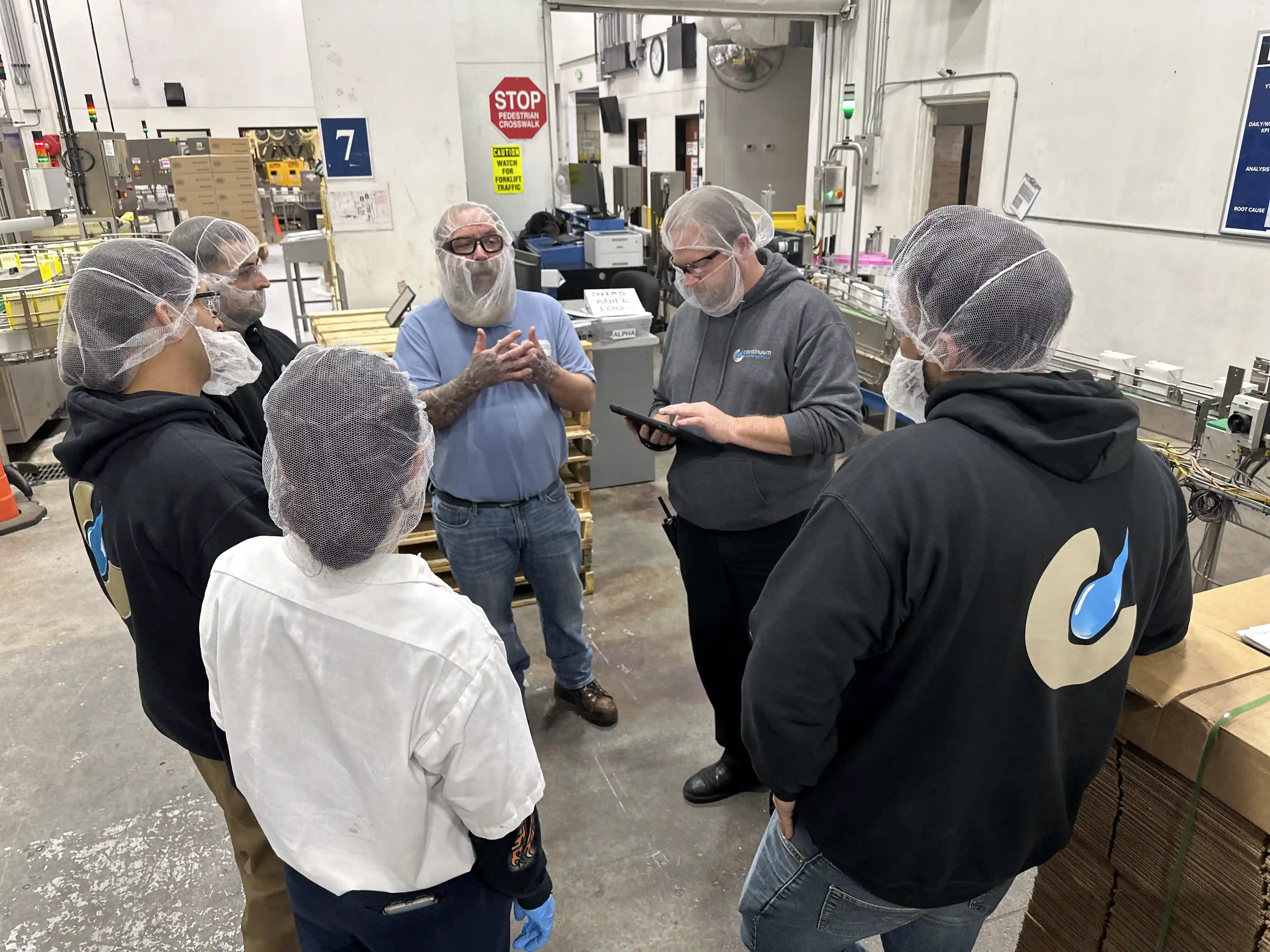 Six factory workers wearing hairnets having a discussion in a manufacturing facility.