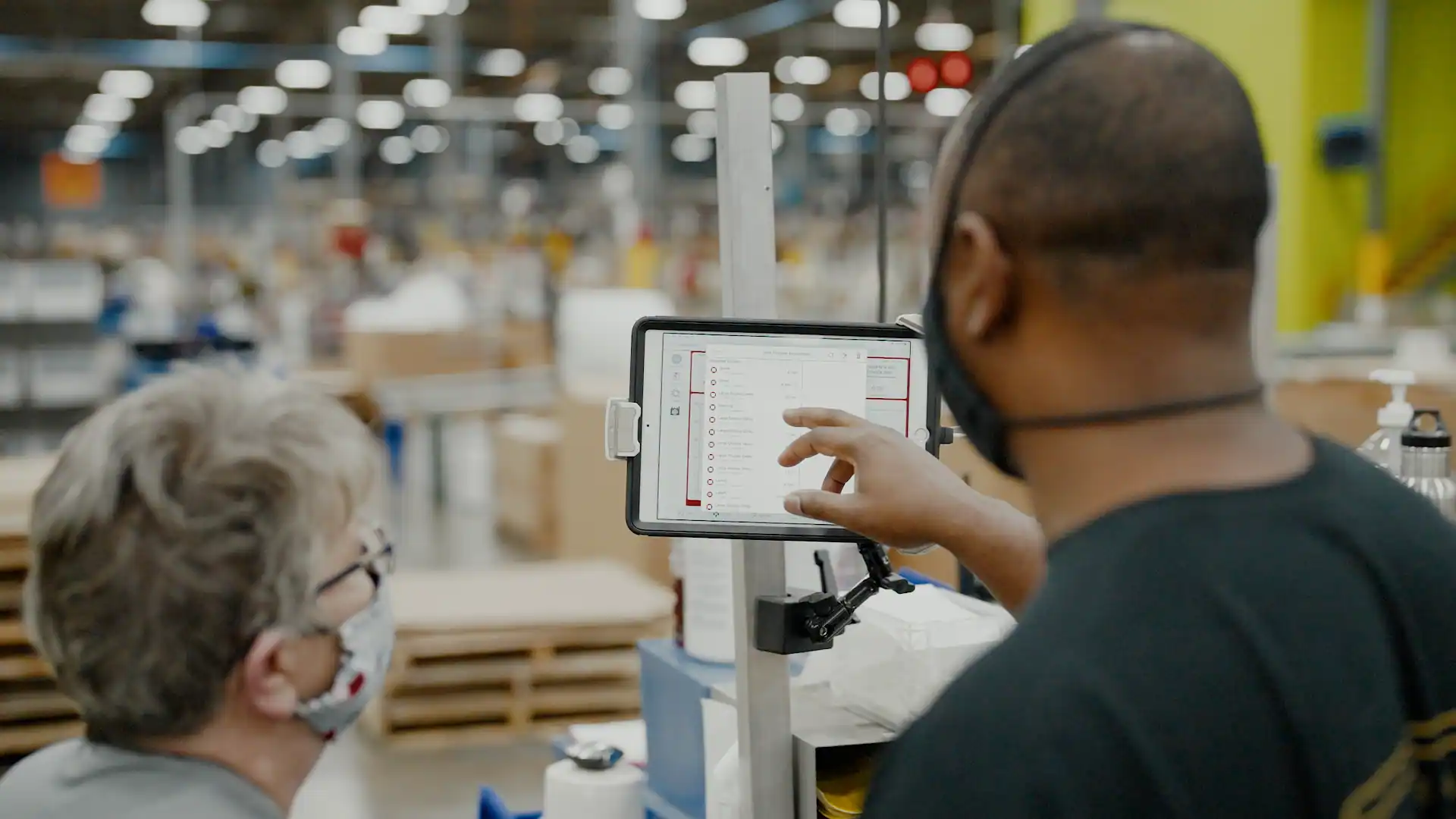 Two masked workers in a warehouse reviewing information on a tablet mounted on a stand.
