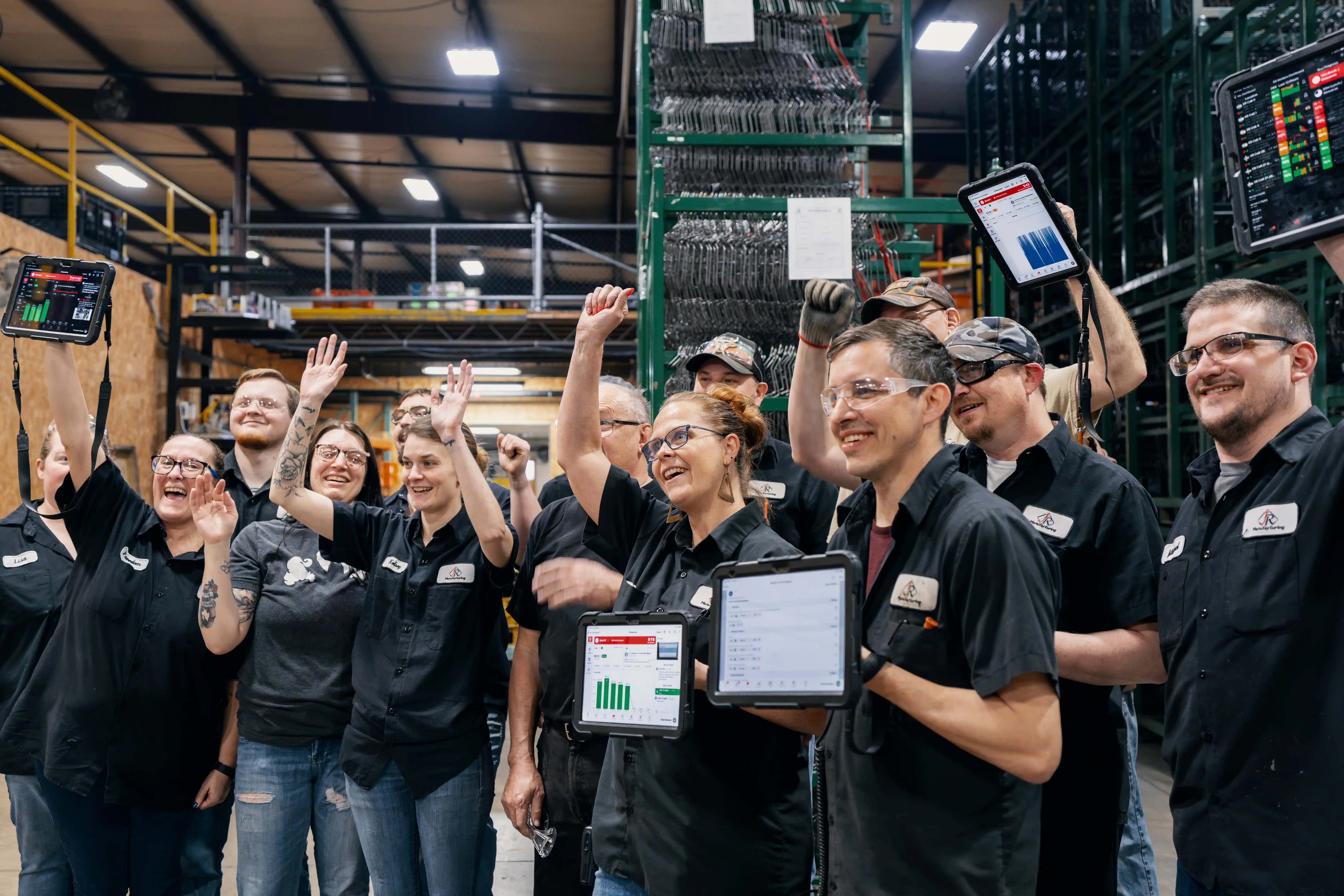 A group of factory workers cheering and holding digital tablets with performance charts in an industrial warehouse.