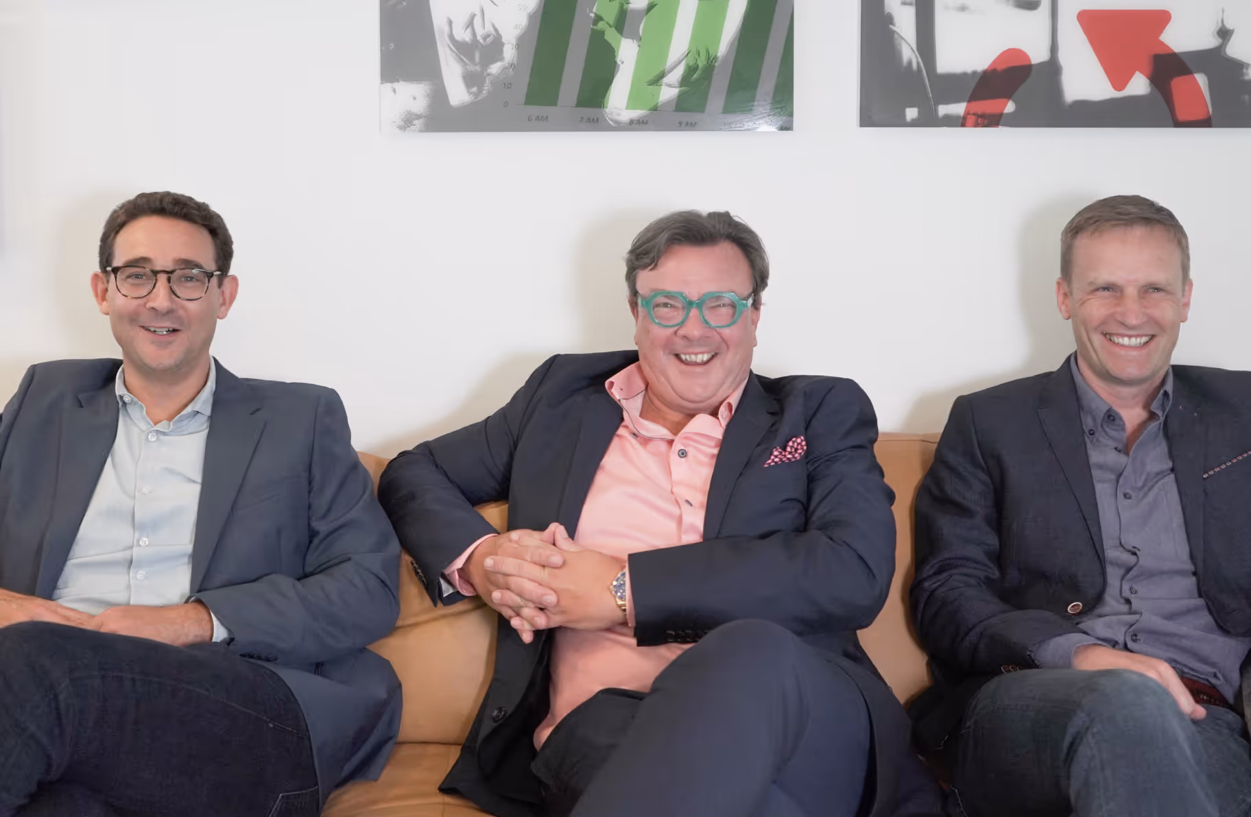 Trois hommes en tenue décontractée souriant assis sur un canapé marron clair contre un mur blanc avec des œuvres d'art encadrées au-dessus.