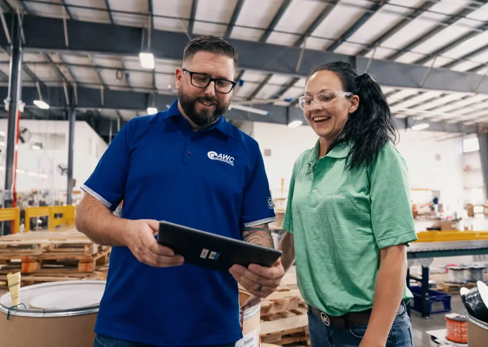 Two workers in a warehouse, a man in a blue AWC shirt holding a tablet and a woman in a green shirt with safety glasses, smiling.