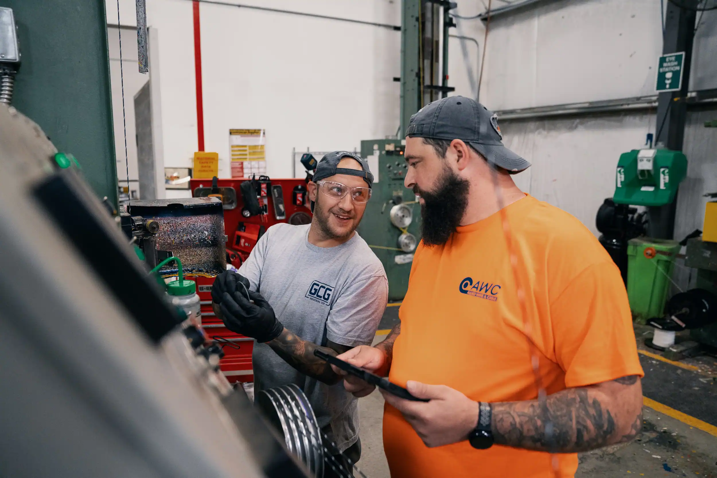Two factory workers in safety glasses and caps discussing work by machinery, one holding a tablet.