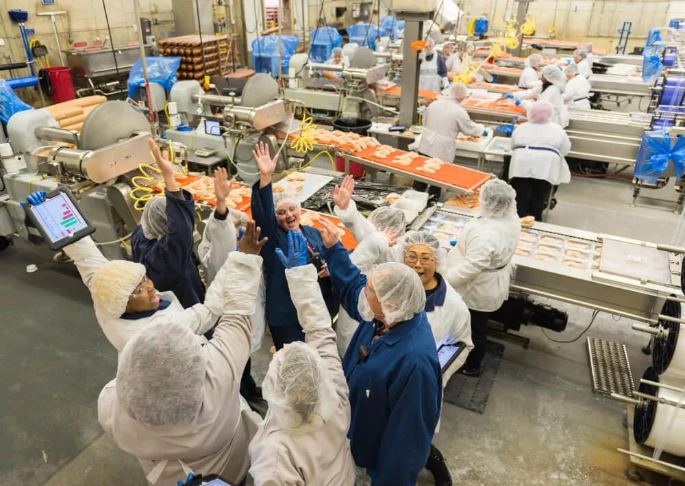 Group of food processing workers wearing hairnets and lab coats giving high-fives in a factory with food conveyor belts.