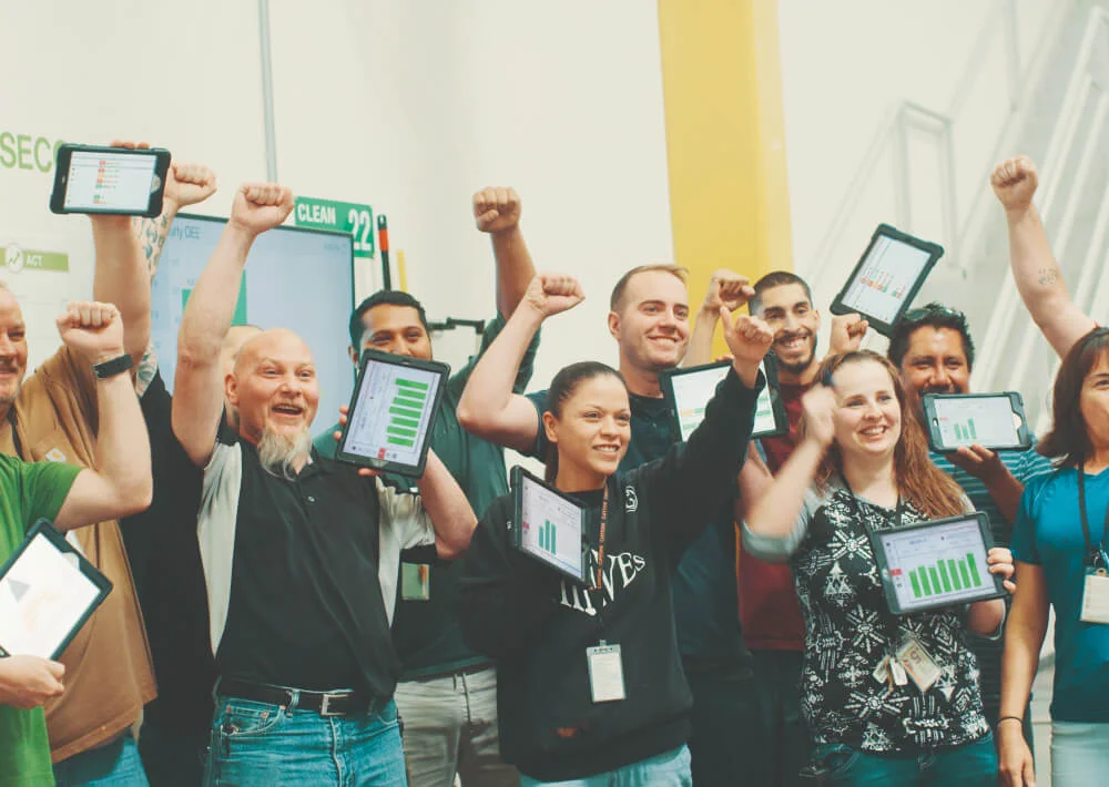Group of diverse coworkers cheering and holding tablets displaying green bar charts in an office setting.
