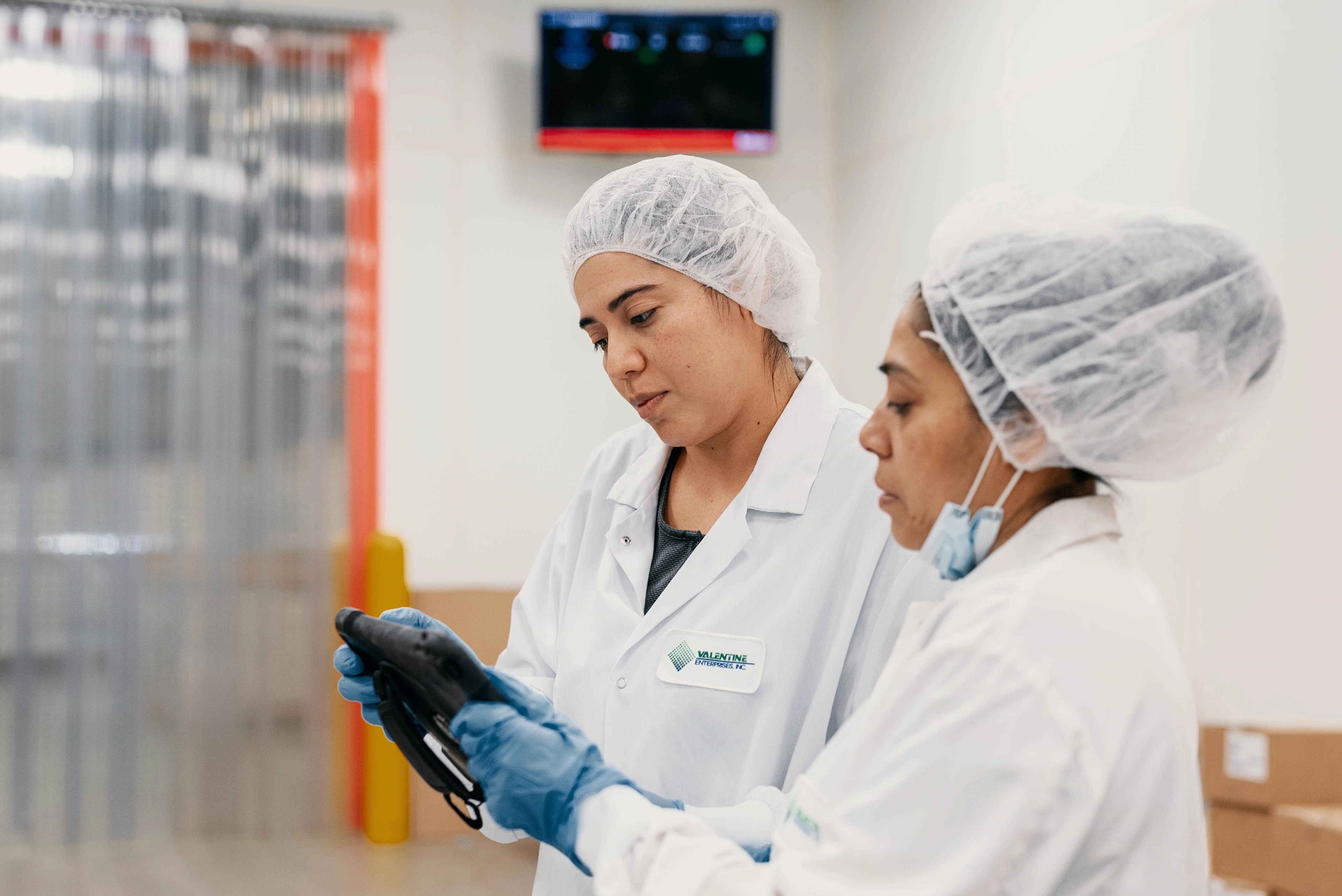 Two female workers in white lab coats, hairnets, and gloves looking at a tablet device in a clean industrial setting.