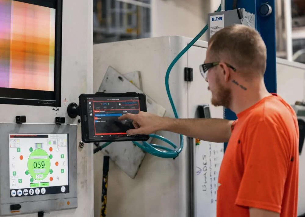 Man wearing safety glasses and an orange shirt operating and pointing at a touchscreen panel in an industrial setting.