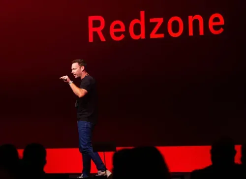 Man in black shirt and jeans speaking on stage with the word 'Redzone' in large red letters behind him.