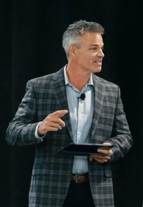 Man in a gray plaid blazer speaking while holding a tablet against a black background.