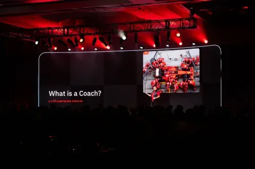Speaker on stage presenting a slide titled 'What is a Coach?' with an image of a Formula 1 pit crew servicing a race car.