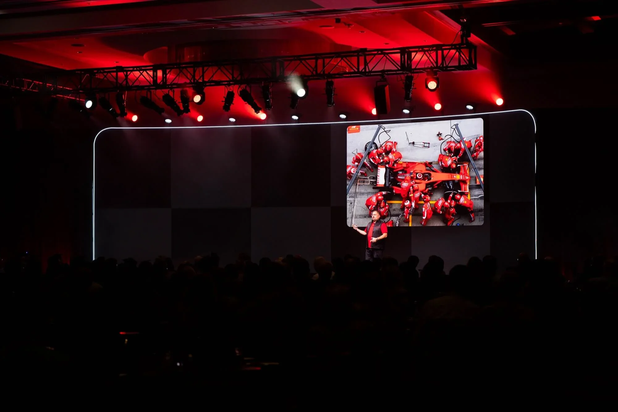 Presenter on stage with a large screen showing a pit stop scene of a red race car and crew.