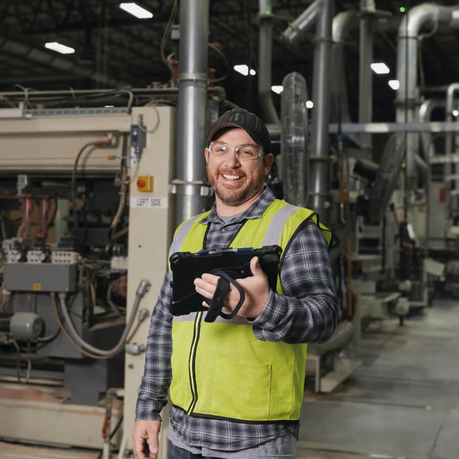 Smiling factory worker in safety vest holding tablet inside industrial facility with machinery.