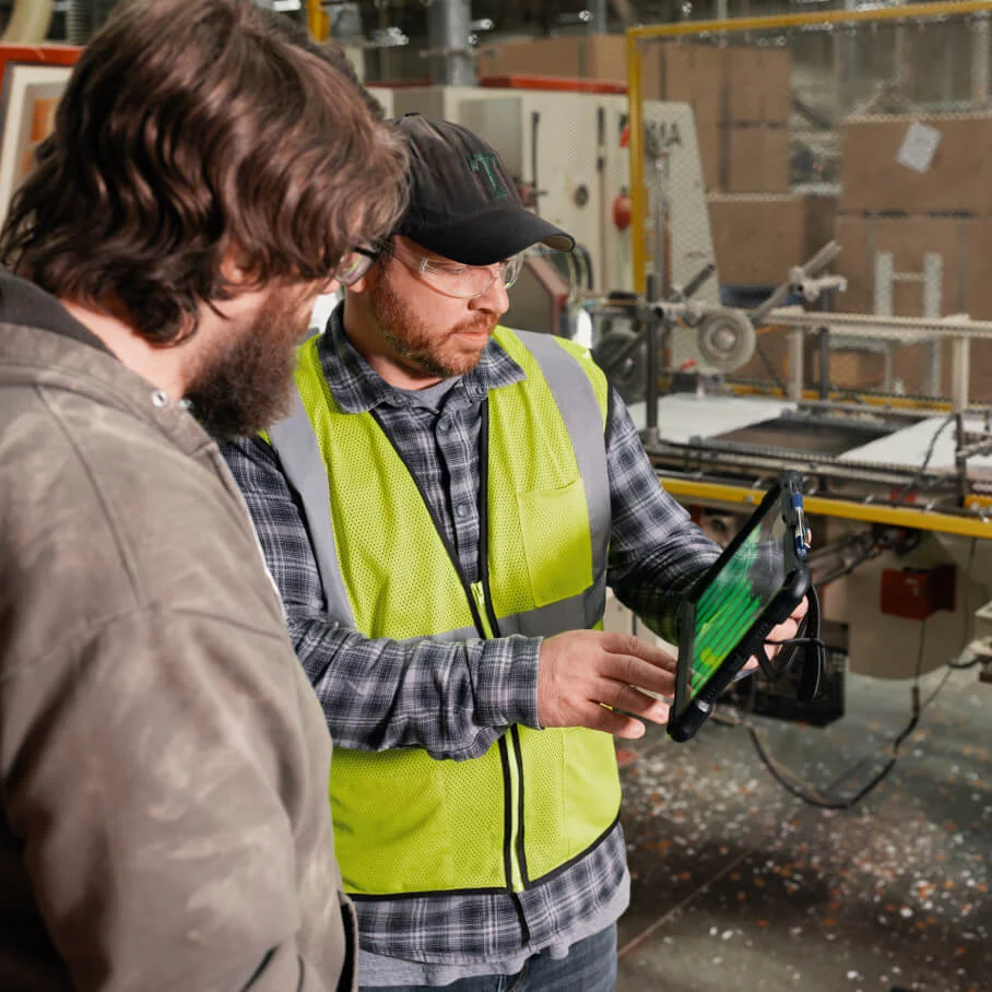 Two workers in a factory, one wearing a safety vest and glasses, using a tablet device.
